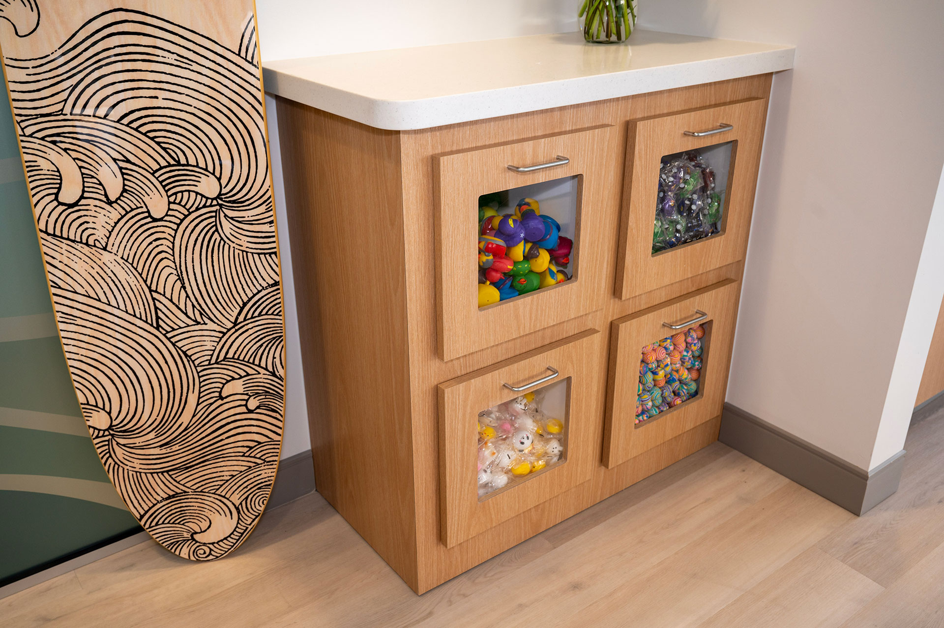 The image shows a kitchen counter with a wooden cabinet featuring multiple drawers, each containing various items such as toys and candies. To the left of the counter stands a surfboard with a unique design on its surface.