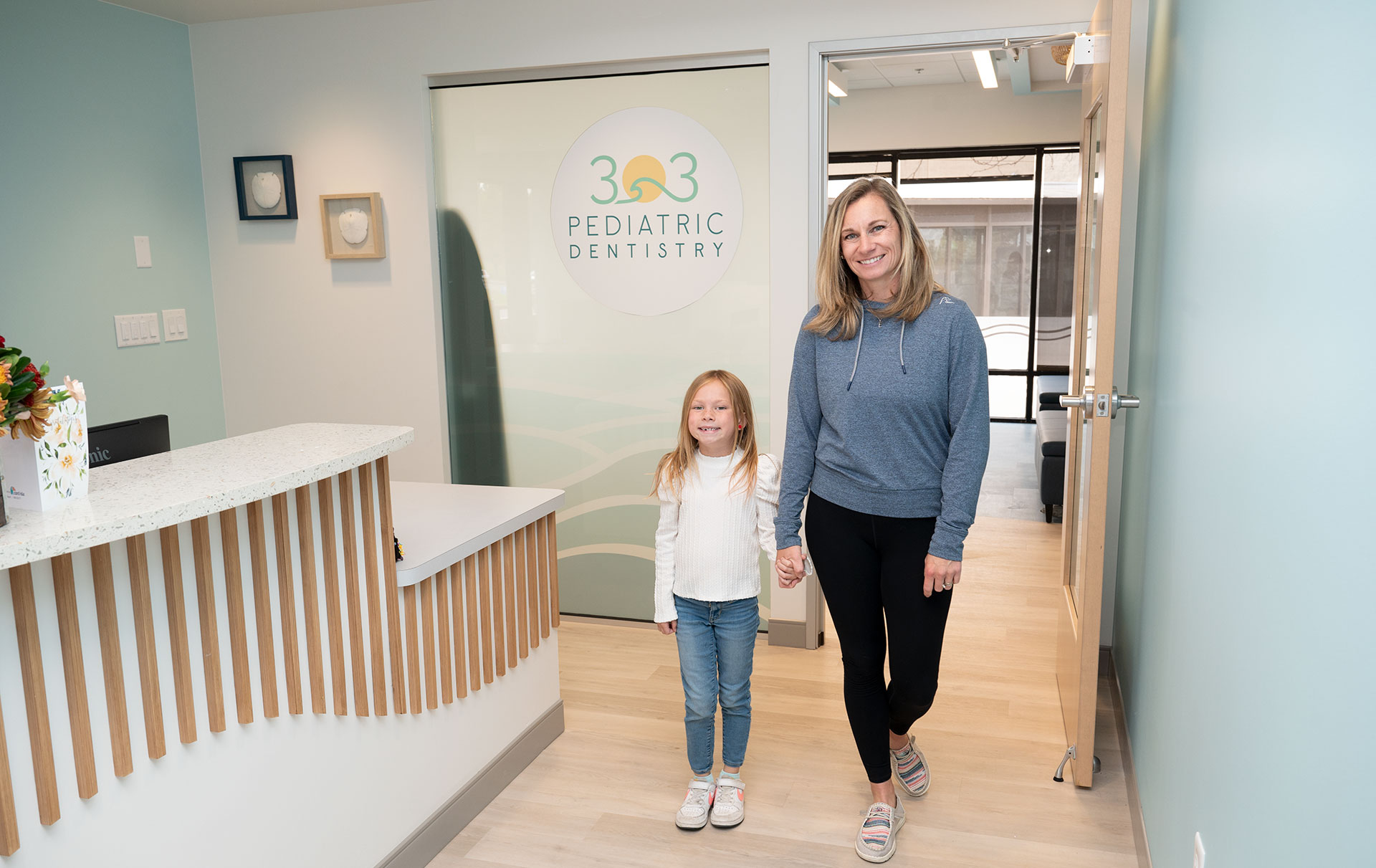 A woman and child walking into a dental office with a sign reading 303 Pediatric Dentistry.