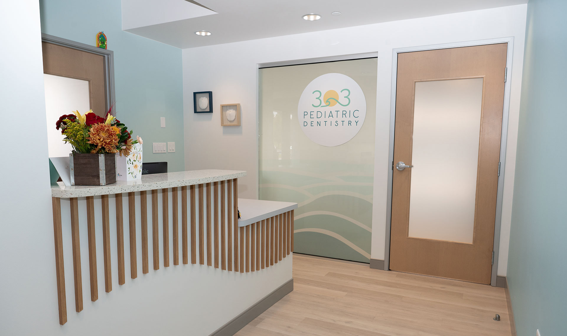 The image depicts an interior view of a dental office reception area with a welcoming atmosphere, featuring a reception desk, a seating area, and a glass door with frosted windows.