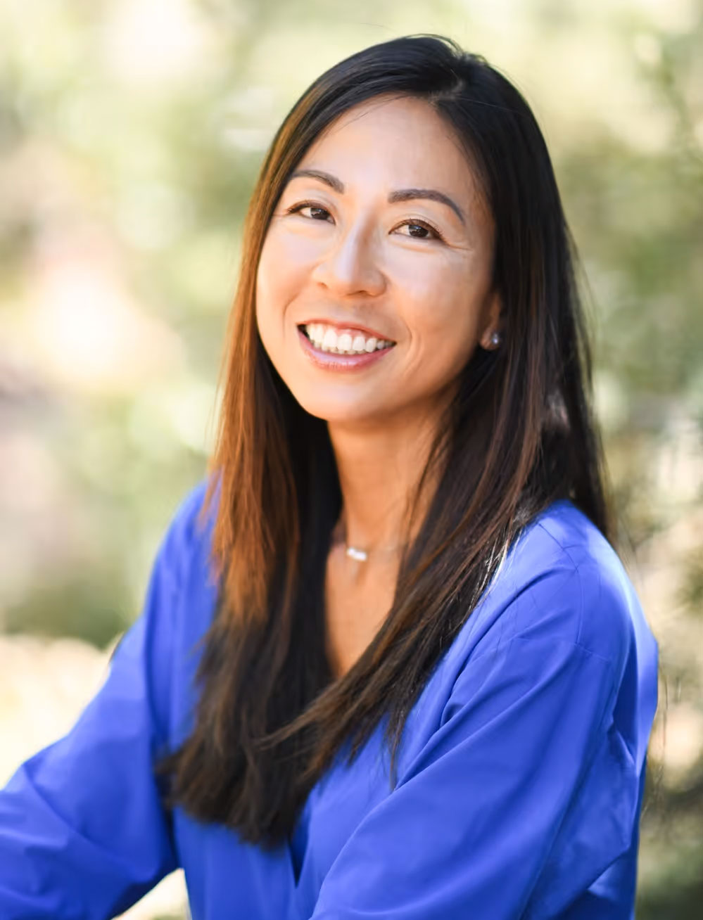 Woman with long dark hair smiling at camera, wearing a blue top and standing outdoors.