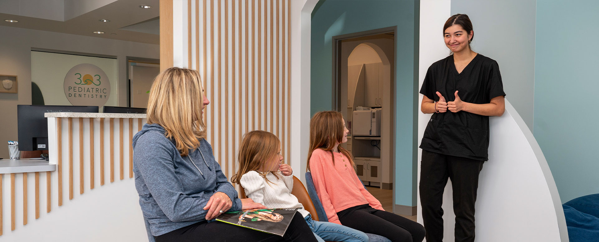 A woman in a black shirt stands with four children in a modern, light-filled room with a curved wall and a blue floor, smiling and gesturing towards them.